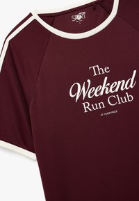 Bordowa koszulka sportowa z teksturowanej tkaniny, białymi akcentami wykończeniowymi oraz centralnym nadrukiem "Weekendowy Klub Biegowy" i "W TWOIM WŁASNYM TEMPIE."