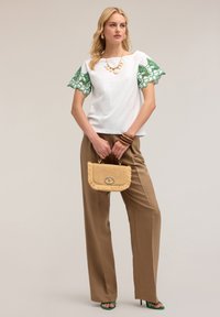 Top bianco con maniche in pizzo verde, pantaloni ampi marroni, borsa di paglia con chiusura dorata e sandali verdi con tacco.