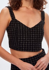 Top court noir avec un décolleté en V, fabriqué dans un matériau texturé présentant un motif à quadrillage blanc. Bretelles épaisses et silhouette ajustée.