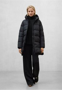 Žena kráčející v černé péřovce s kapucí, černém roláku, kalhotách širokého střihu a černých teniskách na jednobarevném světlém pozadí.