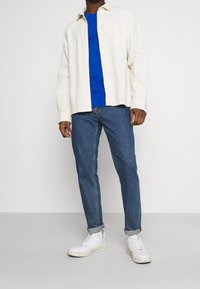 Hellbeige Knopfhemd über einem blauen T-Shirt, kombiniert mit hochgekrempelten dunkelblauen Jeans und weißen Sneakern. Lässiges, geschichtetes Outfit.