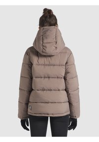 Chaqueta tipo puff en marrón claro, con capucha, diseño acolchado y detalle del logo en el dobladillo. Llevada con guantes y pantalones negros.