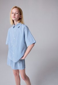 Plava i bijela prugasta košulja na kopčanje s kratkim rukavima, uparena s odgovarajućim šorcima od sličnog materijala. Glatka tekstura, ležerni kroj.