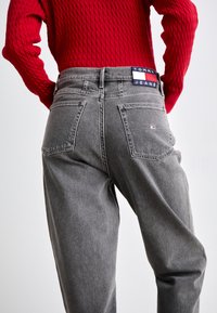 Jeans de mezclilla gris con un ajuste relajado, que cuentan con dos bolsillos traseros y una etiqueta de Tommy Jeans en la cintura. Suéter de punto texturizado rojo visible.