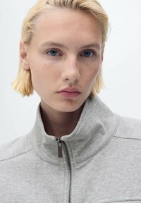 Sweatshirt zippé gris avec un col montant, présentant une texture lisse et un design minimaliste. Le mannequin a des cheveux courts blonds et des yeux bleus.