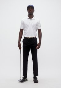 Άνδρας που στέκεται κρατώντας γκολφ ρόπαλο, φοράει λευκό πόλο, μαύρο παντελόνι, μαύρα παπούτσια και μπλε καπέλο, μπροστά σε απλό λευκό φόντο.