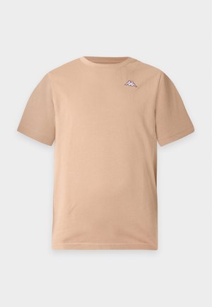Młody mężczyzna z krótkimi włosami, ubrany w beżowy t-shirt z krótkim rękawem z małym logo na piersi oraz oliwkowe spodnie, stojący na jednolitym tle.