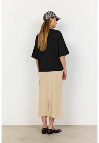 Sort oversized t-shirt, beige midi-skjørt med sidelommer og sorte flade sko, kompletteret med en leopardmønstret kasket, set fra ryggen.