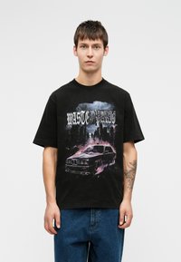 Mladic nosi crnu majicu s grafikom vintage automobila i gradskim obzorjem u ljubičastim nijansama, uparen s plavim trapericama, stoji naslonjen na bijelu pozadinu.