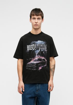 Tânăr îmbrăcat cu un tricou grafic negru, cu o mașină vintage și un orizont de oraș în nuanțe de purpuriu, asortat cu blugi, stând în fața unui fundal alb.