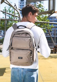 Beige Rucksack mit mehreren Reißverschlusstaschen, gepolsterten Trägern und rechteckiger Form, auf dem Rücken einer Person vor einer Outdoor-Hintergrund platziert.