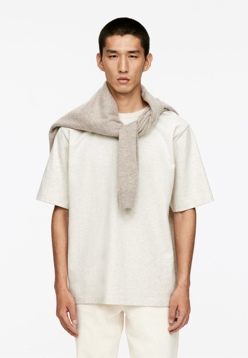 Jeune homme aux cheveux courts portant une chemise beige à manches courtes et un pull léger marron drapé sur ses épaules, sur fond blanc.