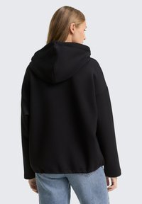 Zwarte hoodie met een relaxte pasvorm, voorzien van een textuurdoek en een geribbelde zoom. Gecombineerd met lichtblauwe denim jeans.