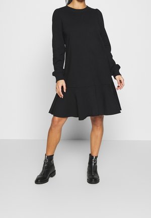 Kobieta w czarnej sukience z długim rękawem, rozszerzanej u dołu oraz w czarnych botkach z zamkami z przodu, stojąca na jednolitym tle.