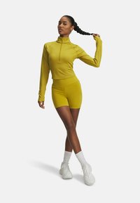 Langsleeve gele sporttop met een rits aan de voorkant, gecombineerd met bijpassende shorts, gedragen met witte sneakers en enkelkousen.