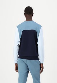 Sudadera con bloques de color en azul marino y azul claro, con cuello redondo, puños acanalados y una textura suave. Presenta un diseño curvado en la parte posterior.