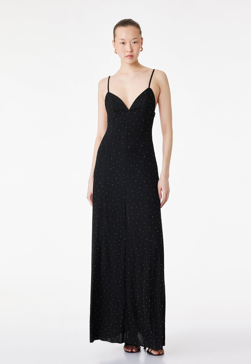 Donna che indossa un lungo vestito nero con spalline sottili e piccoli puntini bianchi, in piedi davanti a uno sfondo bianco uniforme.