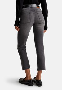 Pantalones de mezclilla gris con pierna recta y acortada, y dobladillo deshilachado. Presenta una cinturilla de altura media y dos bolsillos traseros. Combinados con zapatos negros.