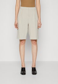 Beige szövetnadrág, egyenes szabással, redőzött elöl, övöltökkel, könnyű anyagból készült. Fekete slip-on cipővel viselve.