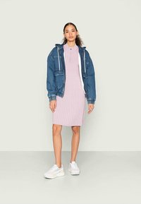 Casaco de ganga com capuz sobre um vestido leve rosa em renda, combinado com ténis brancos. O conjunto tem um corte descontraído e detalhes mínimos.