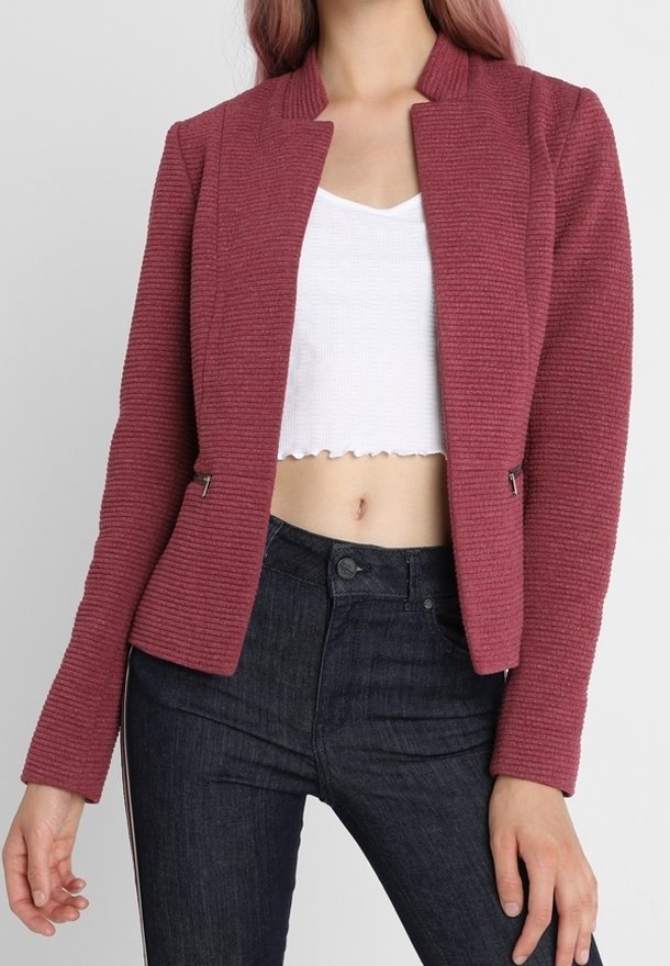 Femme portant un blazer structuré bordeaux sur un top blanc court et un jean taille haute foncé, debout devant un fond uni.