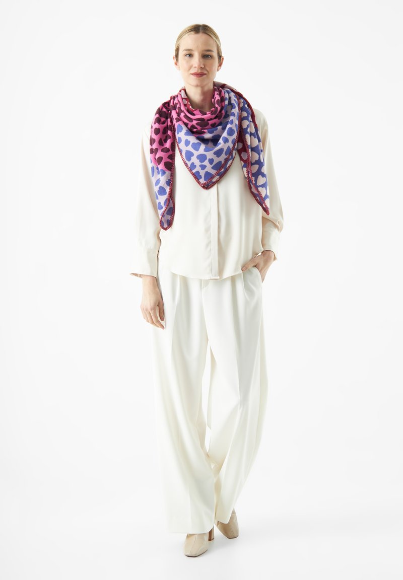 Écharpe en soie rose et violette avec des motifs de cœurs, drapée autour du cou. Modèle portant une blouse blanche et un pantalon blanc ample.