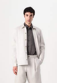 Lys grå button-up jakke med krave, båret over en mørk tæppe skjorte, parret med lys grå bukser; glat stof, minimalistisk design.