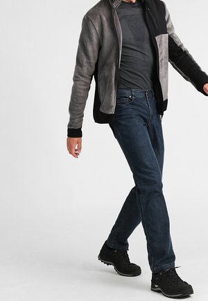 Man loopt en draagt donkerblauwe spijkerbroek, zwarte wandelschoenen, een donkergrijs overhemd en een tweekleurige grijs-zwarte fleecejas.