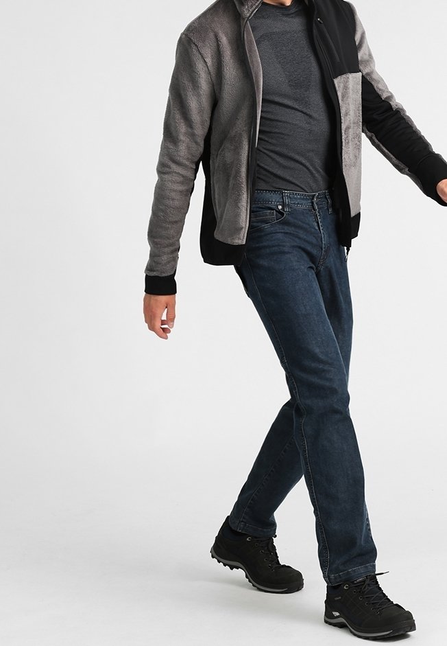 Homme marchant portant un jean bleu foncé, des chaussures de randonnée noires, une chemise gris foncé et une veste polaire bicolore gris et noir.