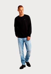 Felpa nera con polsini e scollo a costine, abbinata a jeans azzurri e stivaletti neri alla caviglia. Dettaglio semplice con logo sulla felpa.