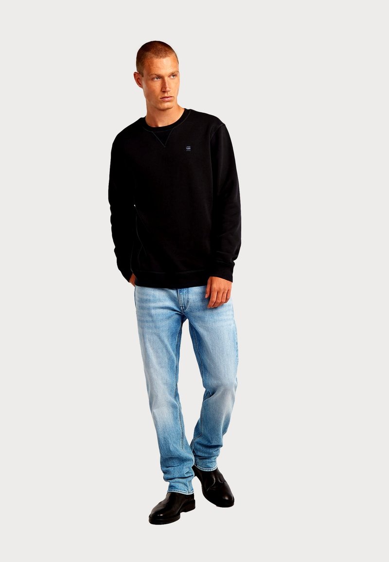 Sweat-shirt noir avec poignets et col côtelés, associé à un jean bleu clair et des bottines noires. Détail du logo simple sur le sweat-shirt.