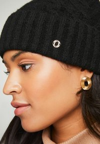 Căciulă din tricot ribbit negru cu un accent logo circular auriu; cercei hoop aurii mari; piele netedă; pulover în nuanțe neutre.