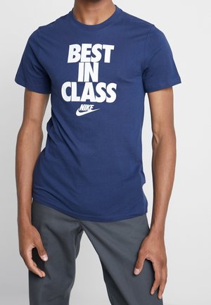 Person trägt ein blaues Nike-T-Shirt mit dem weißen Schriftzug "BEST IN CLASS" und dazu dunkelgraue Hosen vor neutralem Hintergrund.