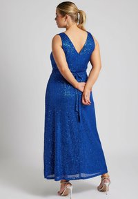 Blå paljettklänning i maxilängd med V-rygg, midjeband och utställd silhuett. Tyget har en strukturerad, skimrande yta. Silvriga klackar synliga.