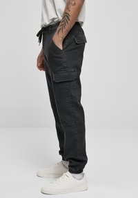 Black cargo pants with side pockets and elastic cuffs. Made of sturdy fabric, featuring a drawstring waist. Worn with white sneakers.