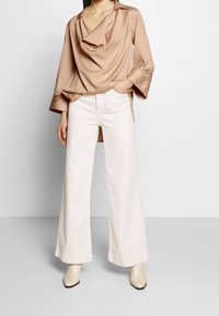 Beige satiinin cowl-neck yläosa pitkähihaisilla hihoilla, yhdistettynä valkoisiin leveälahkeisiin housuihin ja vaalean kremmisiin korokepohjakenkiin.