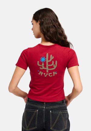 Femme aux longs cheveux foncés porte un t-shirt court rouge avec un dessin de cactus et de soleil, avec le texte « RVCA » au dos, associé à un jean noir.