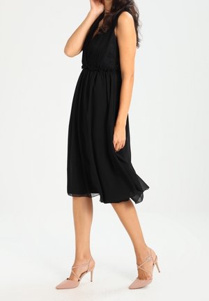 Frau trägt ein ärmelloses schwarzes Midikleid und hautfarbene High Heels, geht und berührt mit einer Hand ihr Haar.