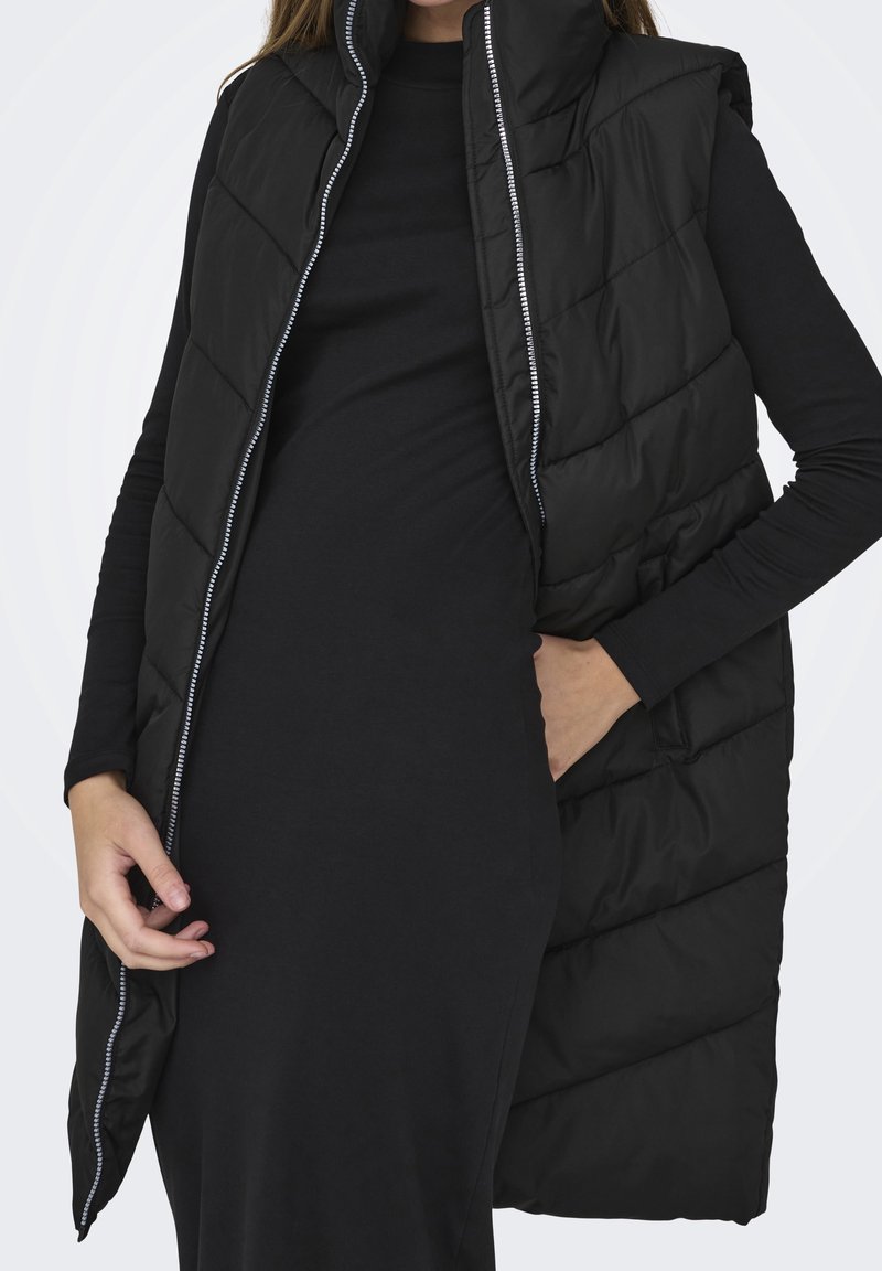 Gilet matelassé noir avec un col montant, présentant un motif matelassé vertical et une fermeture éclair. Porté par-dessus un haut noir à manches longues.