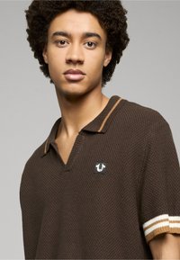 Tricou polo maro tricotat cu decolteu în V, cu accente dungi în bej și alb pe mânecile scurte și un mic logo pe piept.