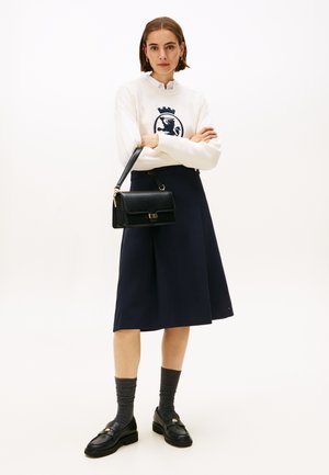 Weißer Pullover mit einem marineblauen Löwenemblem, kombiniert mit einem marineblauen A-Linien-Rock. Schwarze Handtasche mit goldenen Beschlägen und schwarze Slipper mit grauen Socken.