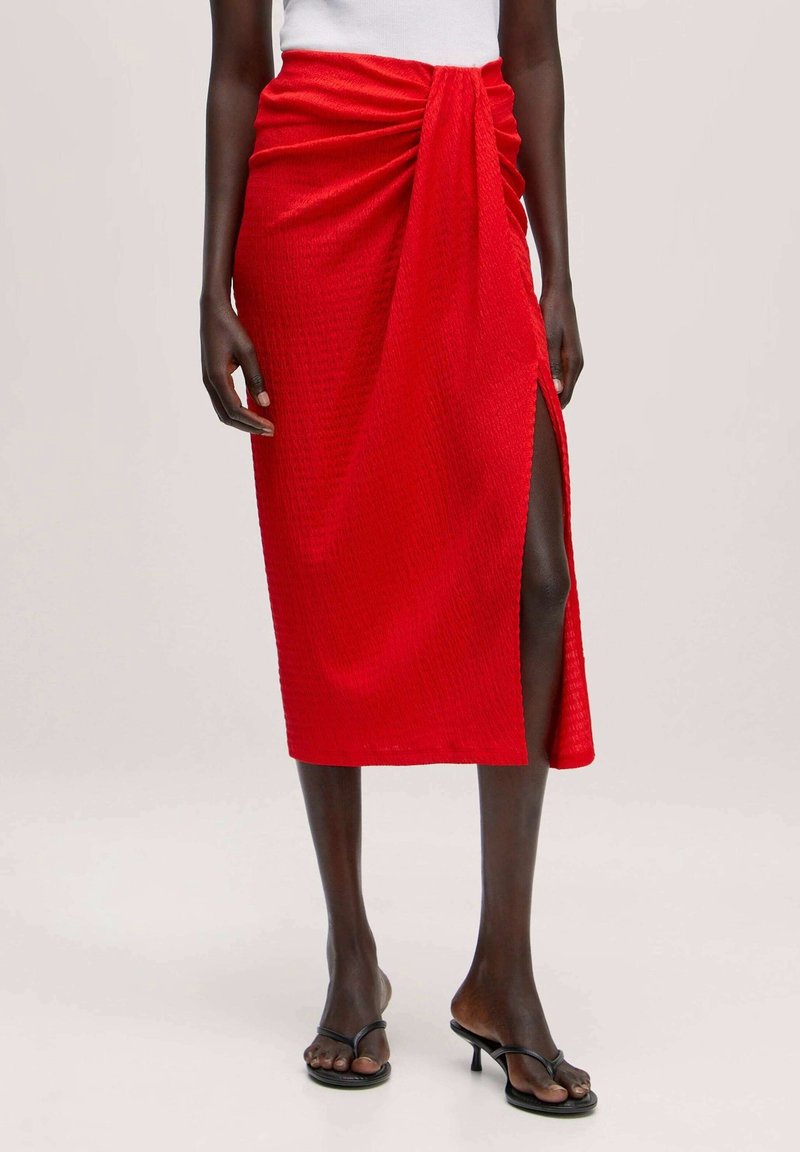 Jupe rouge mi-longue en tissu texturé, avec une fente latérale et des détails froncés à la taille. Associée à des sandales noires à enfiler.