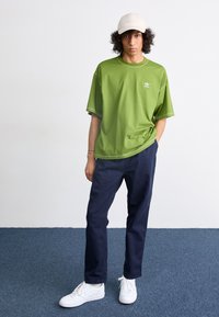 T-shirt surdimensionné vert avec des coutures contrastantes, associé à un pantalon bleu marine et des baskets blanches. Une casquette de couleur claire complète le look.