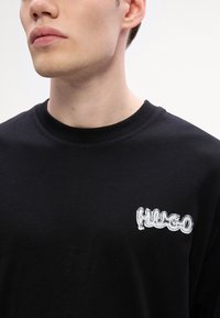 Černé bavlněné tričko s bílým vyšitým logem "HUGO" na levé části hrudi, kulatým výstřihem a krátkými rukávy. Hladká textura.