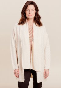 Beige gebreide cardigan met een zachte, gestructureerde afwerking, open voorkant en brede mouwen. Gelaagd over een licht topje en gecombineerd met zwarte broek.