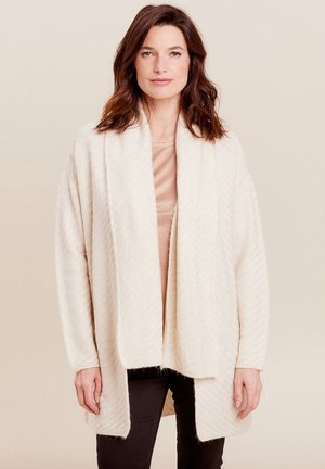 Beige gestrickte Strickjacke mit einer weichen, strukturierten Oberfläche, offener Vorderseite und weiten Ärmeln. Über einem hellen Oberteil getragen und mit schwarzen Hosen kombiniert.
