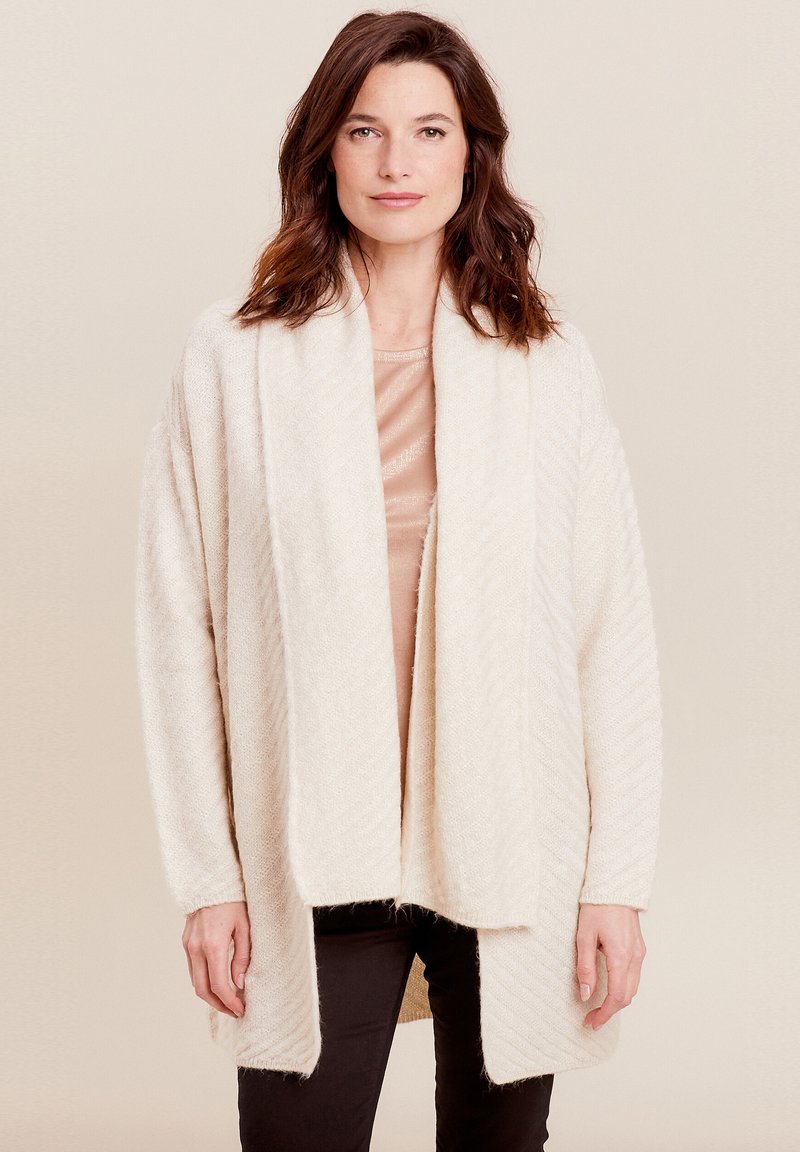 Beige gebreide cardigan met een zachte, gestructureerde afwerking, open voorkant en brede mouwen. Gelaagd over een licht topje en gecombineerd met zwarte broek.