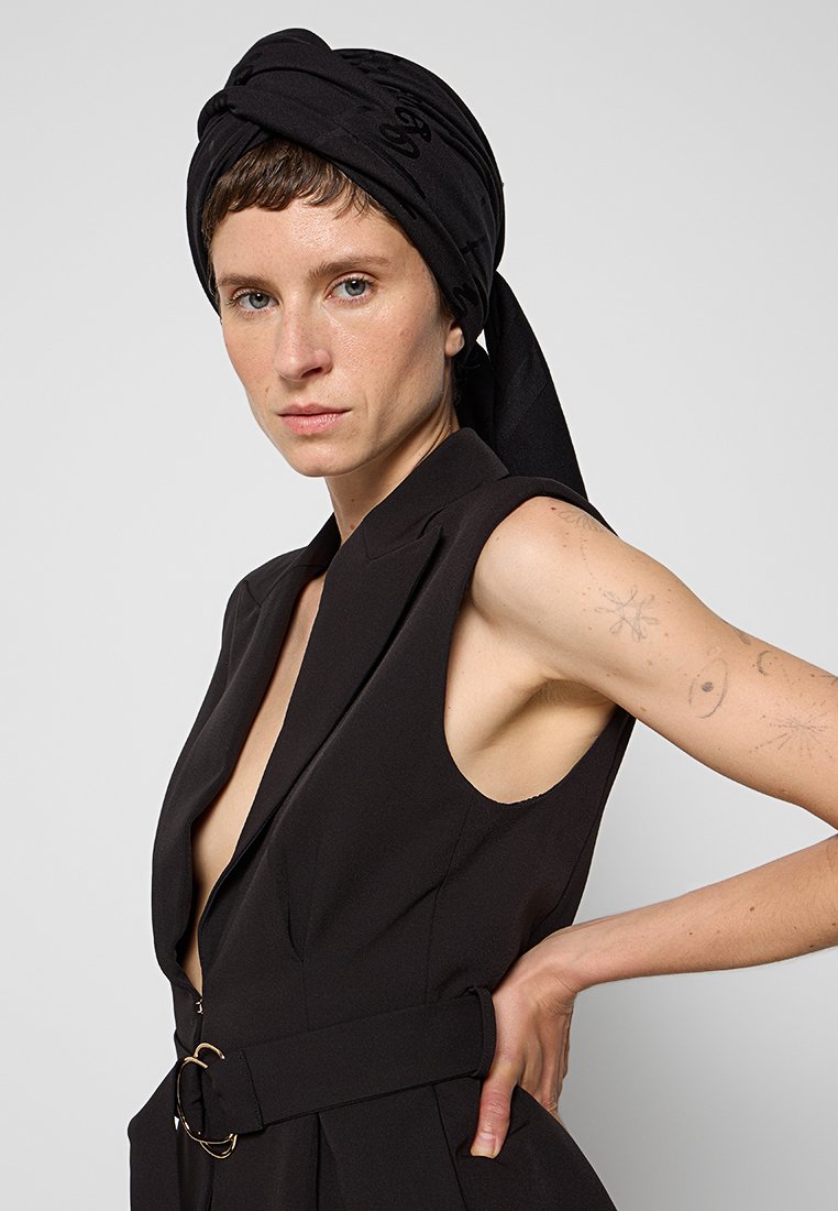 Czarna bezrękawnikowa sukienka o głębokim dekolcie w kształcie litery V, z wcięciem w talii i złotymi akcentami; zestawiona z czapką turbanową.