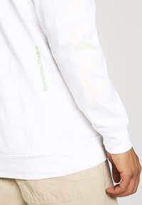 Pitkähihainen valkoinen paita, jossa on vihreitä ja vaaleankeltaisia printtejä hihoissa, valmistettu pehmeästä puuvillasta ja varustettu joustimilla.