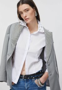 Blazer gris à fines rayures avec des clous argentés sur les épaules, chemise blanche avec col orné, ceinture noire et jean en denim. Bracelets argentés portés.